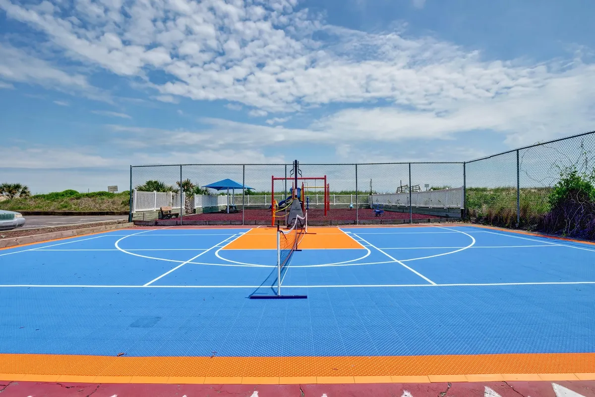 An inviting outdoor sports court with vibrant colors, dual basketball hoops, and shaded spectator seating surrounded by lush greenery. The setting is clearly designed for both basketball and tennis, ideal for friendly competitions and group activities.