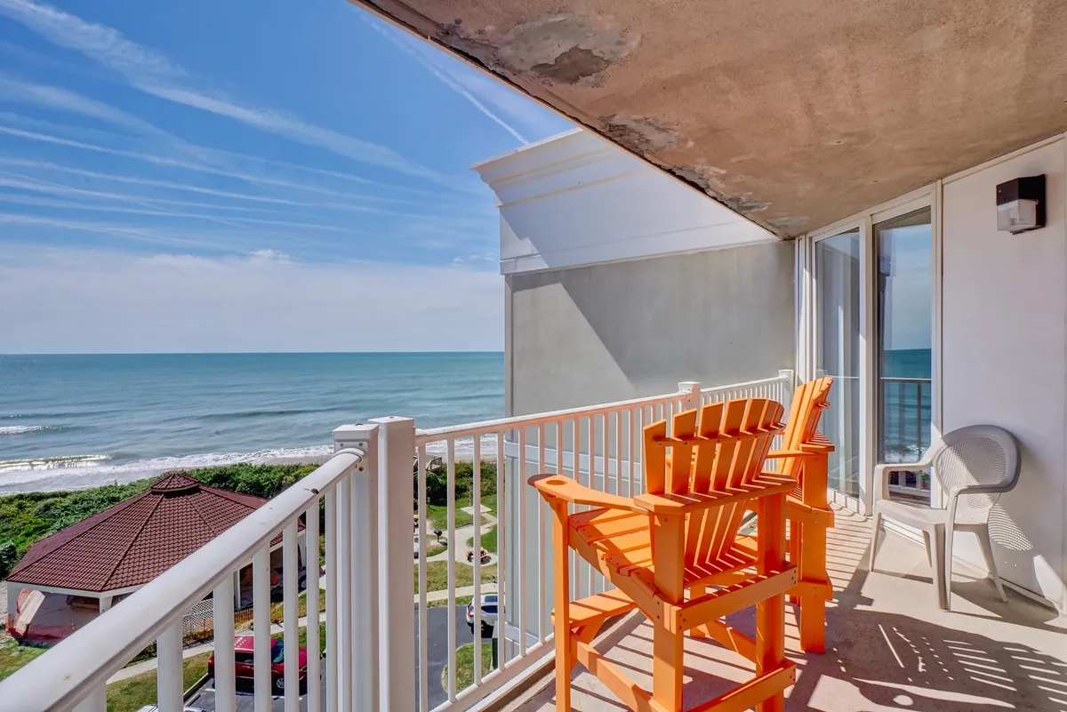 This image features two vibrant orange Adirondack chairs set on a balcony or patio, with a sweeping view of the ocean and a clear blue sky stretching to the horizon. The scene suggests a tranquil, private space perfectly suited for relaxing and enjoying the sound of the surf whether in the early morning or at sunset.