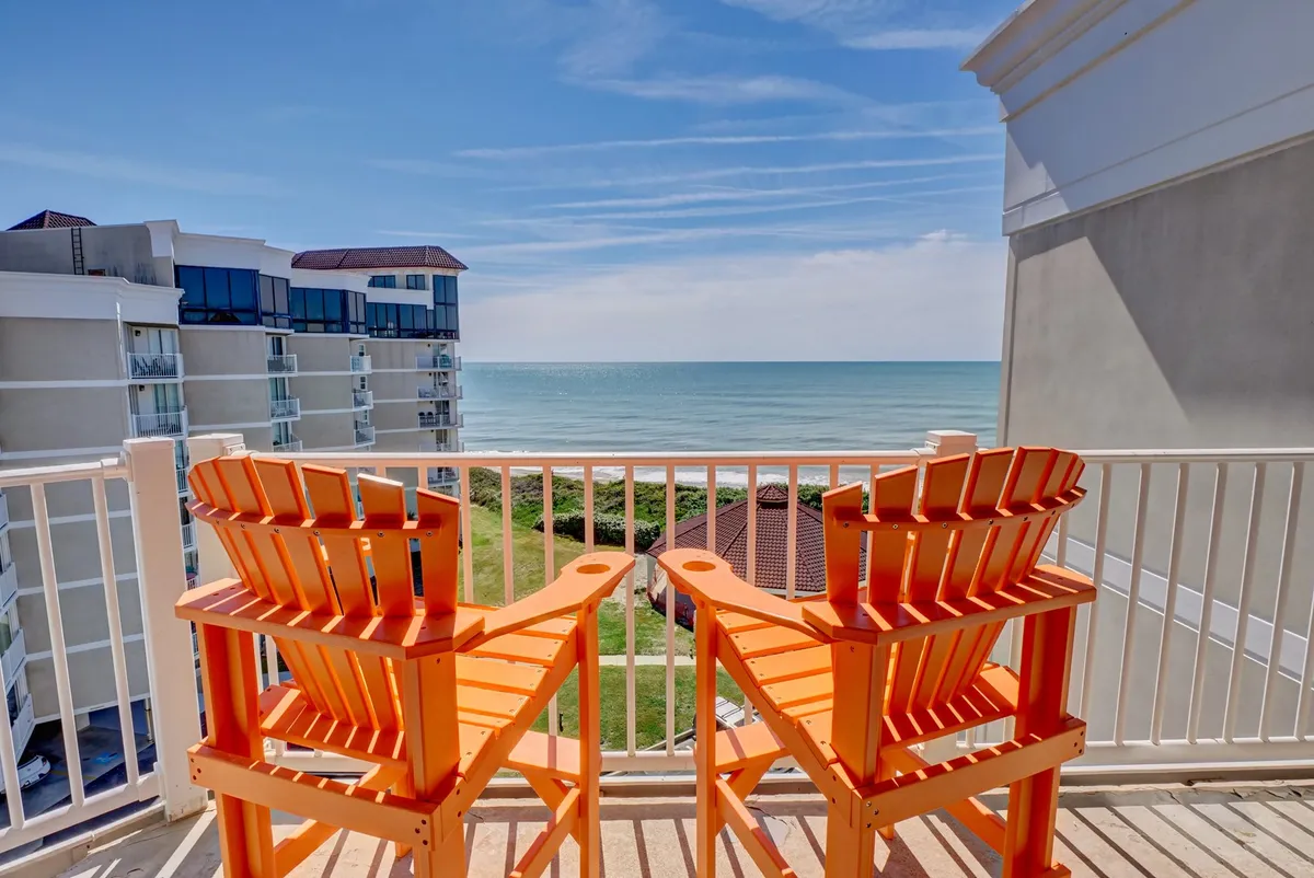 The image features a pair of vibrant orange chairs on a private balcony overlooking the vast, sparkling Atlantic Ocean at North Topsail Beach. The clear blue sky and gentle waves set a relaxing, inviting scene, perfect for unwinding, enjoying sunrise views, or soaking in the soothing ocean breezes.