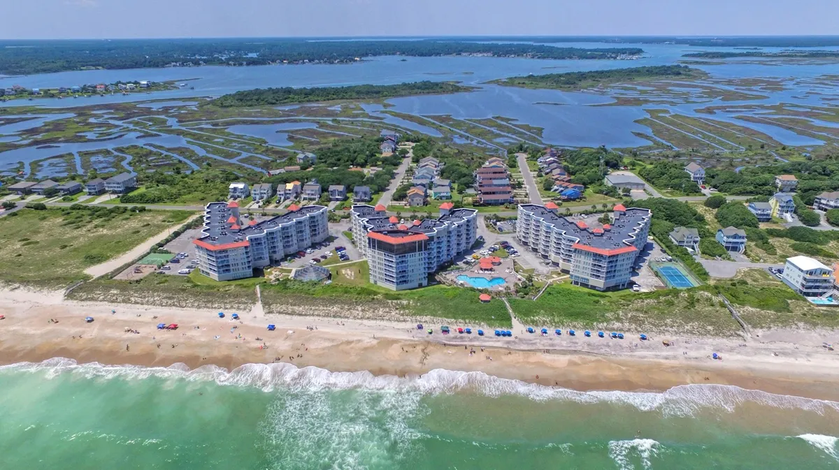 A sweeping view of the beachfront property, showcasing the swimming pool, surrounding marshlands, ocean waves, and colorful beach umbrellas. The scene is lively and captures the essence of a beachside family resort.