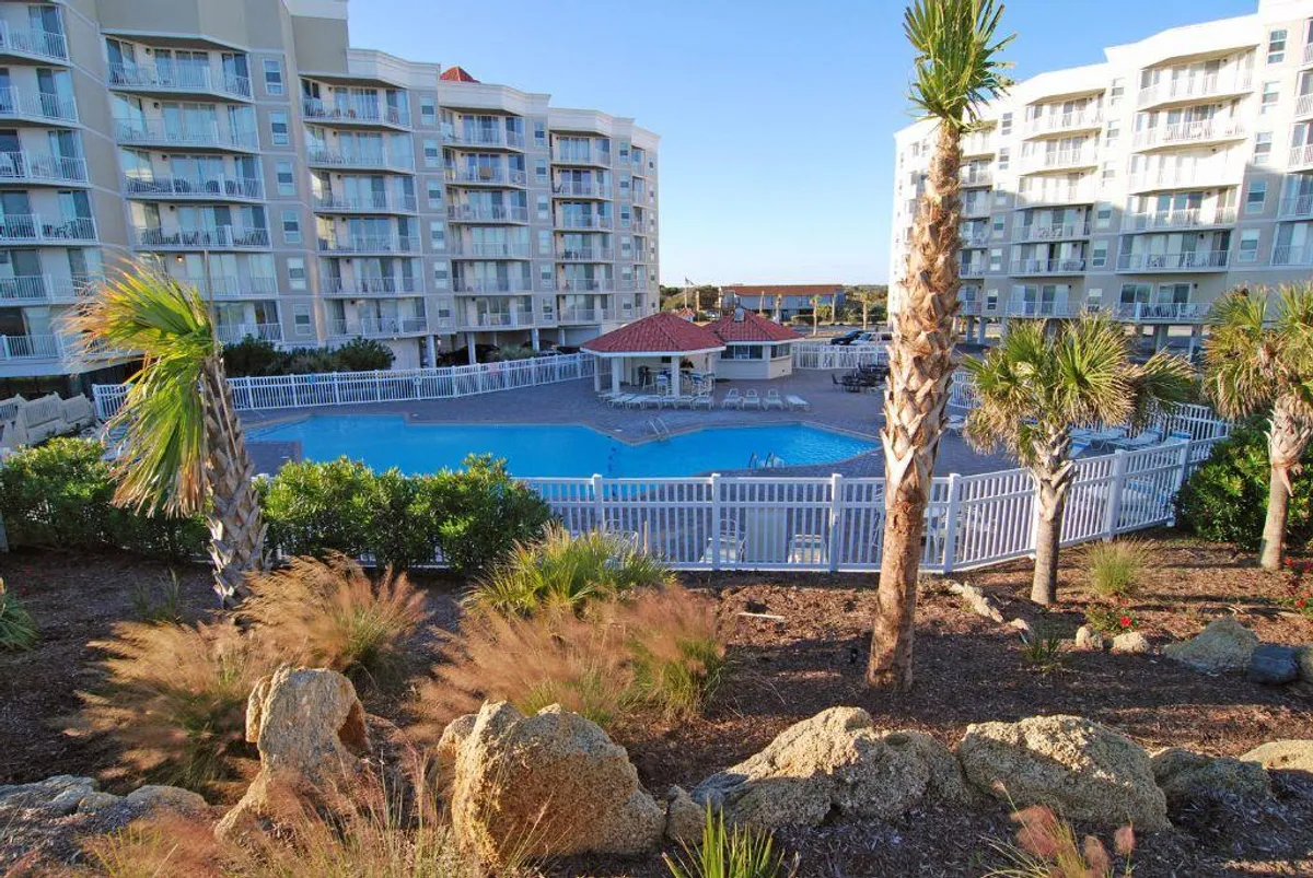 An inviting outdoor swimming pool is surrounded by palm trees and landscaped gardens, with lounging areas and nearby buildings visible. The tranquil setting is perfect for both relaxation and social gatherings.
