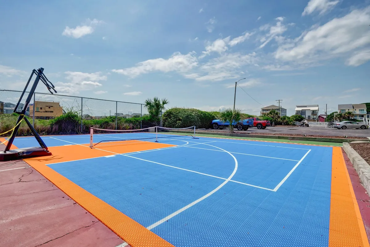 A brightly colored basketball court with a tennis net stretched across, set amidst lush greenery. The court is open and inviting, suggesting opportunities for basketball, tennis, and other friendly outdoor games.