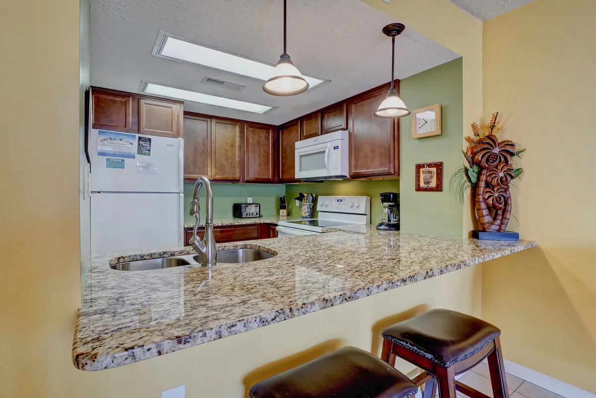 A modern kitchen with granite countertops, wood cabinets, decorative wall art, stainless steel appliances, and bar stools at the counter. The space feels warm and welcoming, ideal for cooking and casual dining.