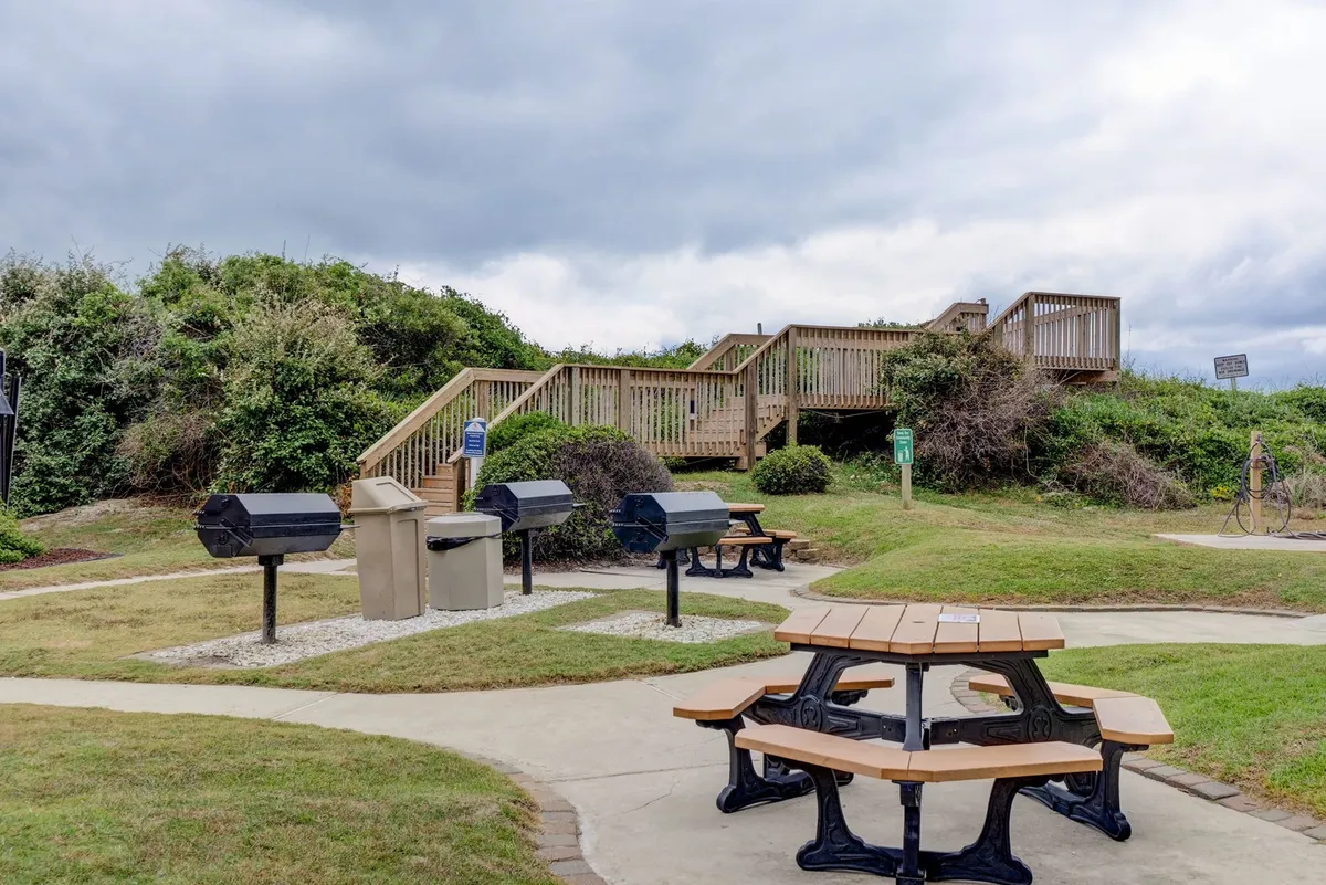 An outdoor grilling and picnic area with several barbecue grills, wooden picnic tables, stairs leading from the building, and trash bins nearby. The relaxed ambiance suggests communal gatherings and easy alfresco dining.