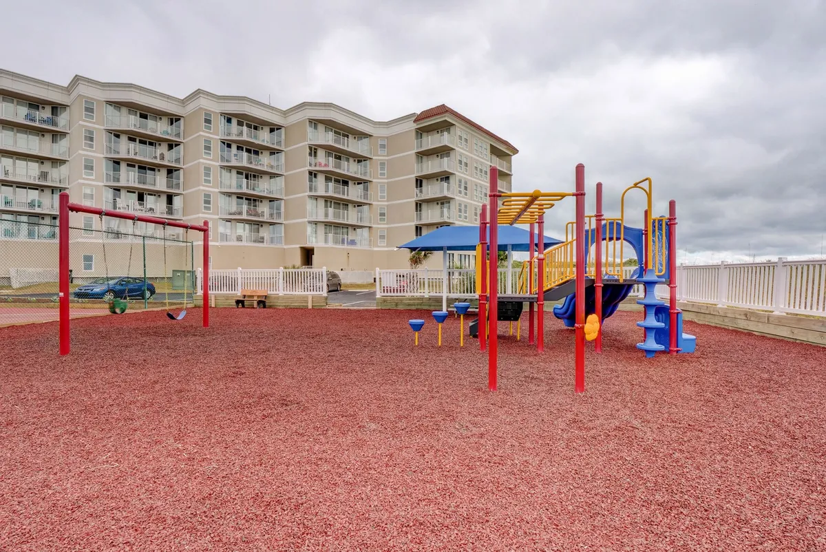 A vibrant playground area featuring modern play equipment, swings, and colorful rubber flooring. The area is surrounded by greenery and invites families and children to gather and interact in a lively, safe environment.