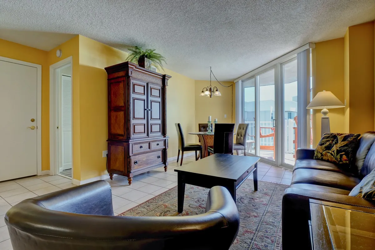 A cozy, sunlit living space featuring a wooden cabinet, dining area for four, a comfortable sofa, and a coffee table. Natural light pours in through large windows, brightening the room and creating an inviting setting for gathering and relaxation.