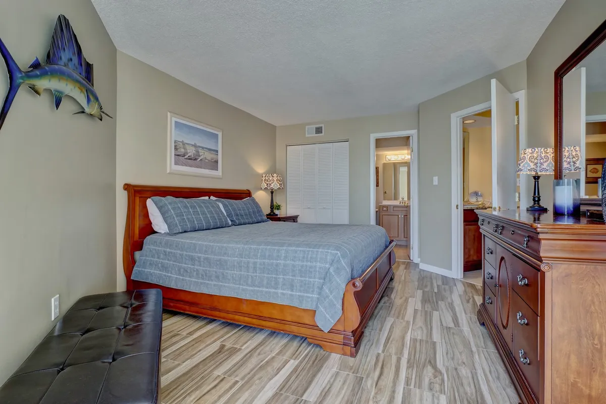 A tranquil primary bedroom featuring a plush king-sized bed, a wooden dresser, and coastal decorative accents like fish wall art. The soft lighting and serene color palette establish a restful atmosphere ideal for unwinding after a day at the beach.