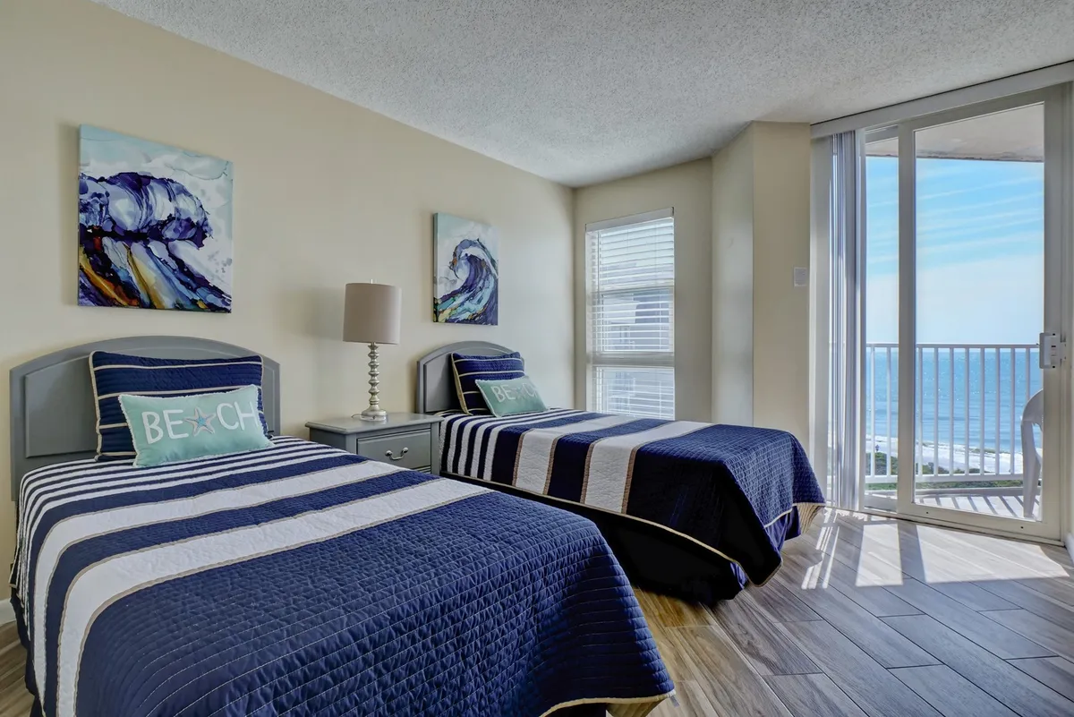 A bright second bedroom with two twin beds dressed in blue and white bedding, decorative beach-themed pillows, and wall art depicting ocean waves. A large sliding glass door leads directly to a private balcony, offering a glimpse of the ocean beyond.